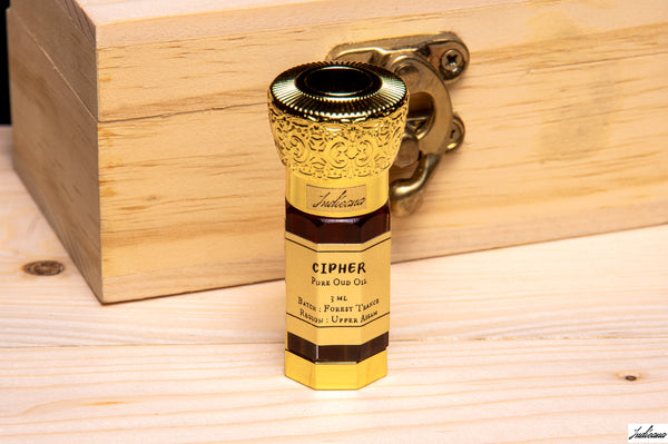 A gold-colored Cipher Oud Oil bottle with a golden cap, standing on a light wooden surface in front of a wooden box.