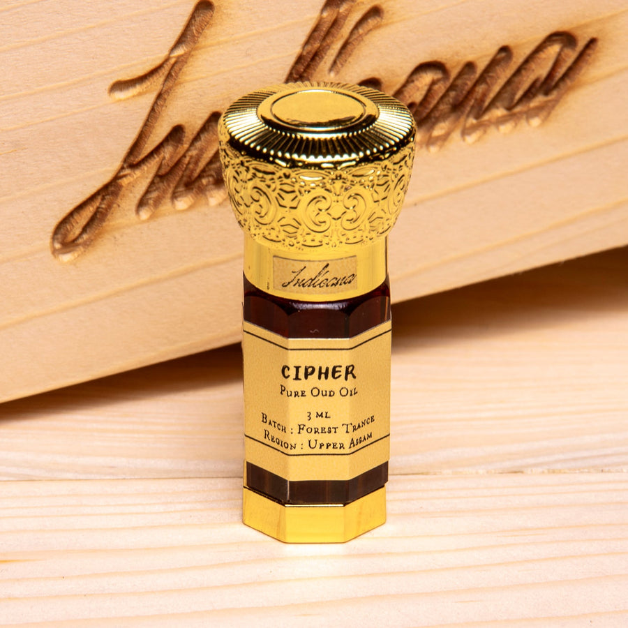 A gold-colored Cipher Oud Oil bottle with a golden cap, standing on a light wooden surface in front of a wooden box.
