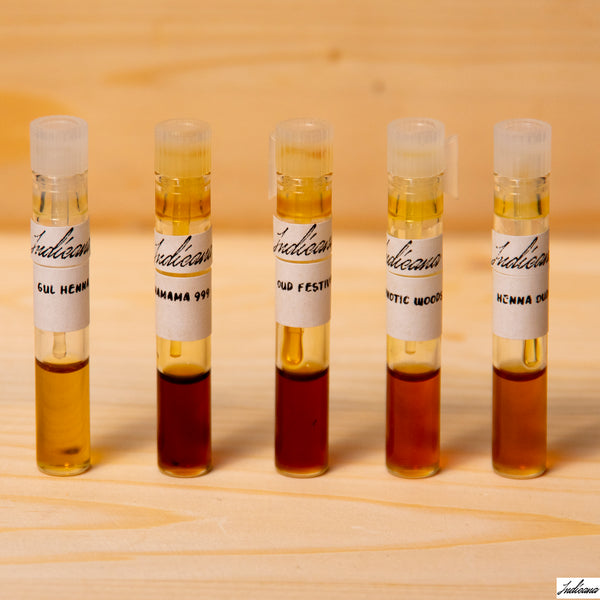 Five small clear vials with white labels containing attars by Indicana Oud, arranged in a row on a light wood surface against a warm beige background.