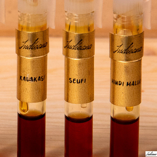 Three small glass vials with gold-colored metal collars labeled 'Indicana' and handwritten names 'KALAKASI', 'SEUFI', and 'HINDI MALiKI' on a wooden surface.