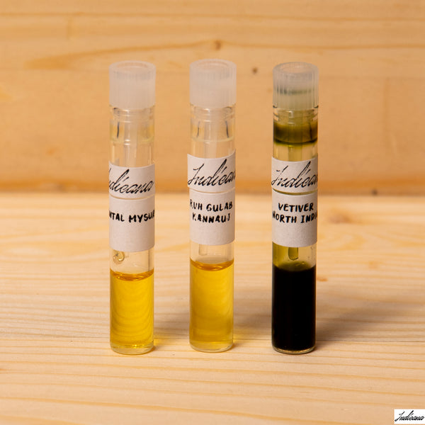 Three small labeled glass vials containing essential oils by Indicana Oud, standing on a light wood surface against a pale wood background.