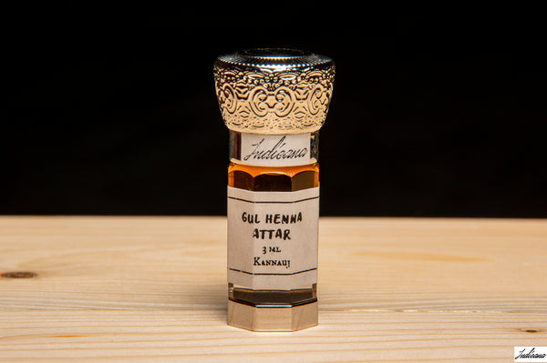 A small glass bottle of Gul Henna Attar perfume oil with a white label and an ornate silver cap, set on a light wooden surface against a dark background.