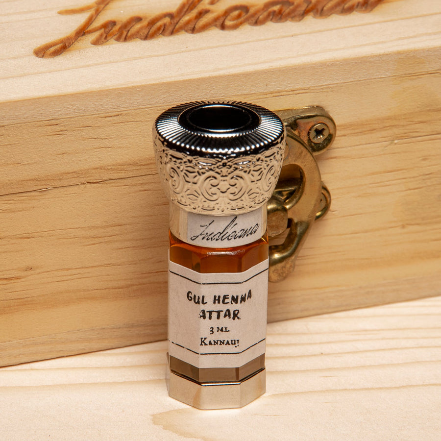 A small glass bottle of Gul Henna Attar perfume oil with a white label and an ornate silver cap, set on a light wooden surface against a dark background.