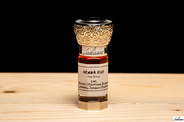 A small glass bottle of Henna Oil with an ornate gold-toned cap, standing on a light wood surface against a solid black background.