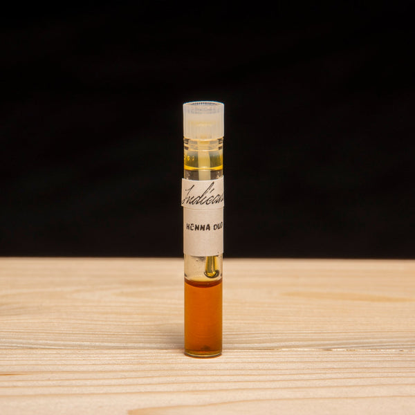 A small glass vial with a white label stands upright on a light wood surface against a black background. The vial contains attar by Indicana Oud.