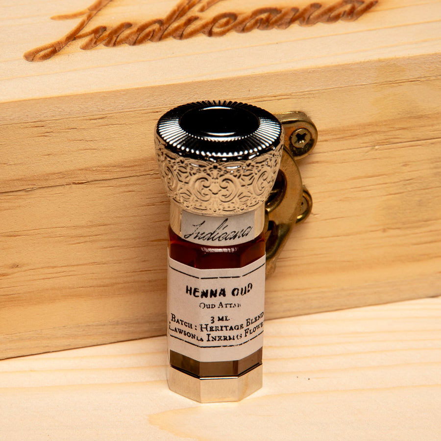 A small glass bottle of Henna Oil with an ornate gold-toned cap, standing on a light wood surface against a solid black background.