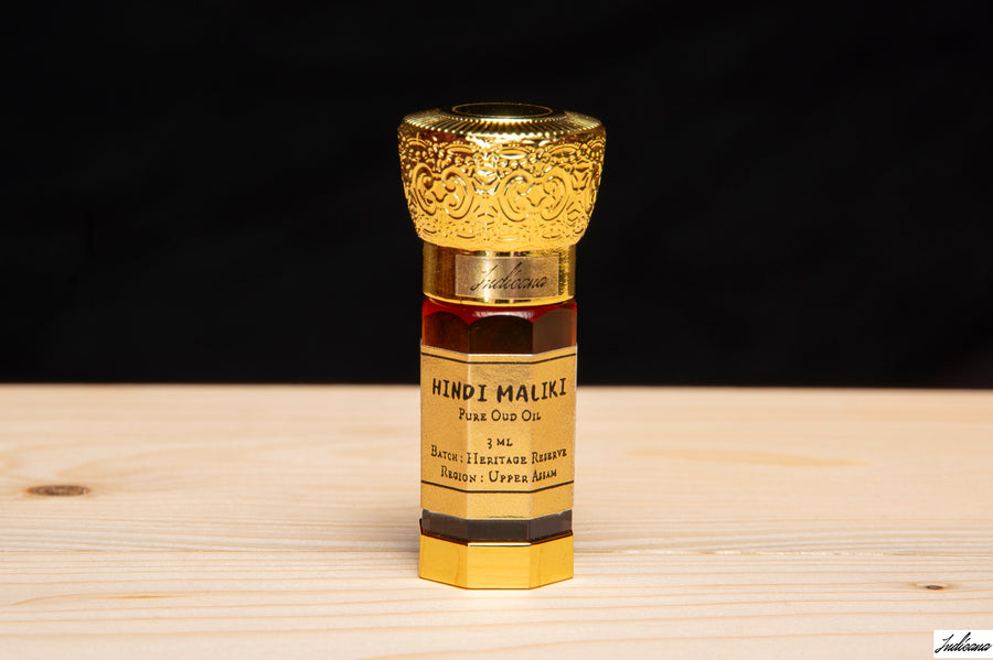 A small bottle of Hindi Maliki Oud Oil stands in front of an open wooden gift box with white paper filler. The bottle has a gold-toned ornate cap, a dark amber liquid, and a cream label with black text.
