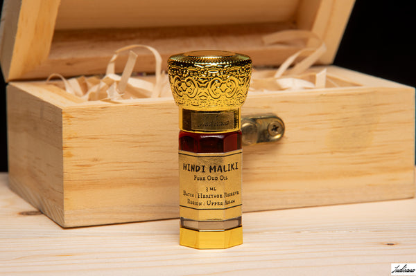 A small bottle of Hindi Maliki Oud Oil stands in front of an open wooden gift box with white paper filler. The bottle has a gold-toned ornate cap, a dark amber liquid, and a cream label with black text.