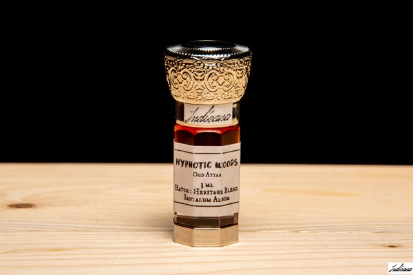 A small glass vial of 'Hypnotic Woods' by Indicana Oud, featuring a decorative silver-toned cap and a white label, set on a light wood surface against a solid black background.