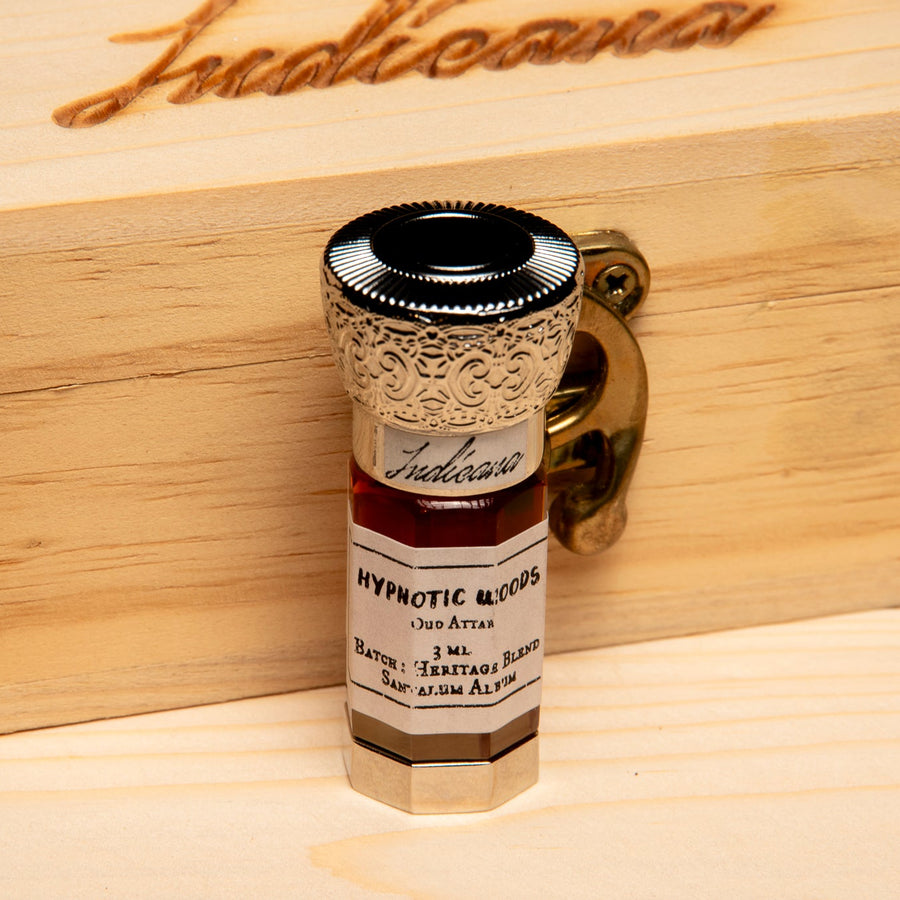 A small glass vial of 'Hypnotic Woods' by Indicana Oud, featuring a decorative silver-toned cap and a white label, set on a light wood surface against a solid black background.