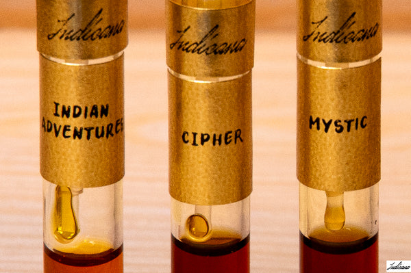 Three small glass vials labeled 'Indicana' with handwritten names 'Indian Adventures,' 'Cipher,' and 'Mystic,' filled with oud oils, shown on a light wood surface.