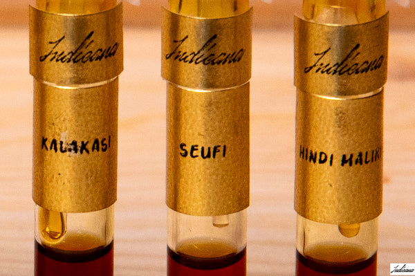 Three small glass vials with gold-colored metal collars labeled 'Indicana' and handwritten names 'KALAKASI', 'SEUFI', and 'HINDI MALiKI' on a wooden surface.
