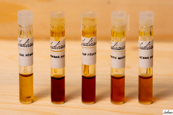 Five small clear vials with white labels containing attars by Indicana Oud, arranged in a row on a light wood surface against a warm beige background.