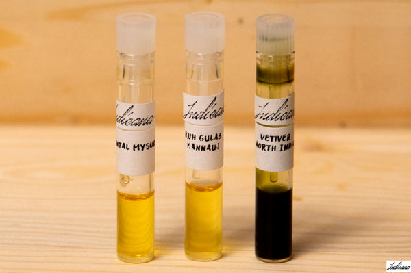 Three small labeled glass vials containing essential oils by Indicana Oud, standing on a light wood surface against a pale wood background.