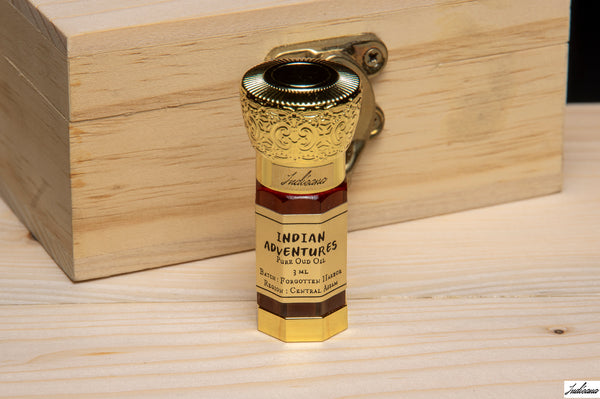 A brass hook lock designed to look like a vintage Indian Adventures Oud Oil box, And the bottle featuring an ornate engraved top and a printed label, shown resting against a light wood box on a wooden surface.