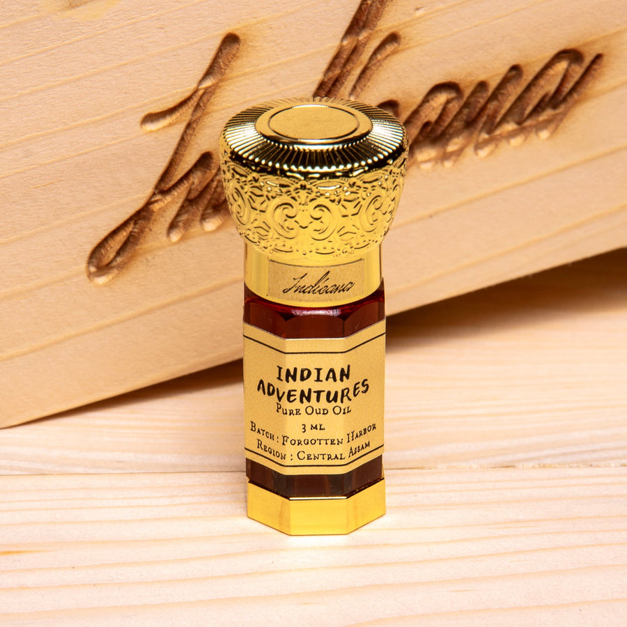 A small bottle of Indian Adventures Oud Oil stands upright on a light wood surface in front of a larger wooden box. The bottle is gold-toned with dark brown label bands and black text, and the background is a natural wood setting.