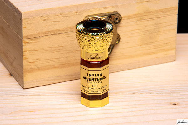 A small bottle of Indian Adventures Oud Oil stands upright on a light wood surface in front of a larger wooden box. The bottle is gold-toned with dark brown label bands and black text, and the background is a natural wood setting.