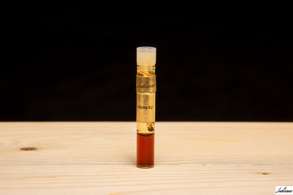 A small cylindrical glass vial with a white cap stands upright on a light wood surface against a dark black background. The vial contains oud oil.