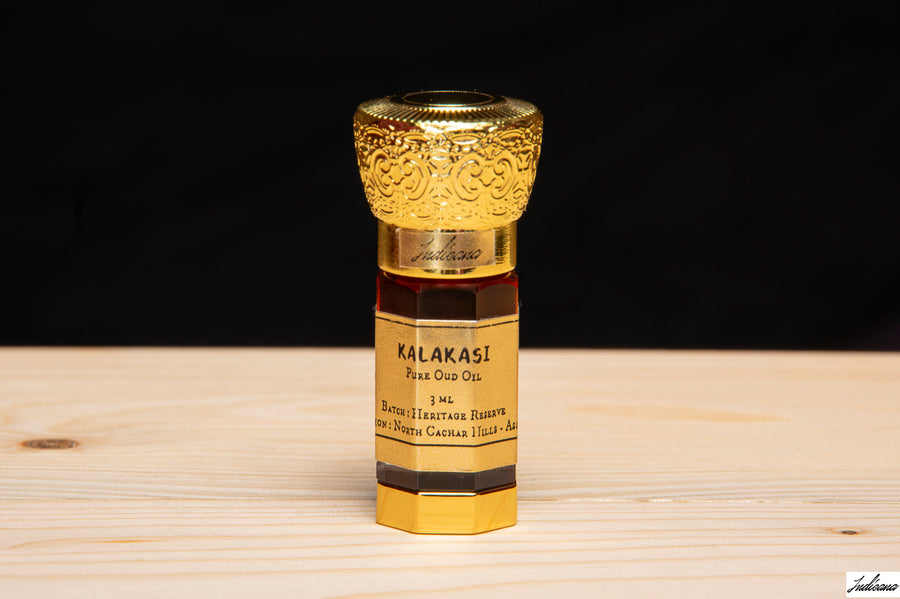 A small bottle of Kalakasi Pure Oud Oil with an ornate gold cap stands in front of an open wooden gift box on a light wood surface.