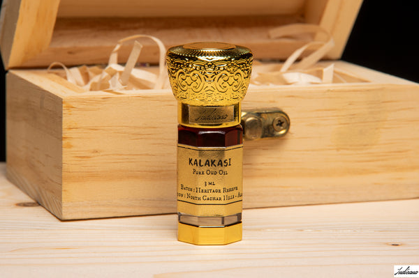 A small bottle of Kalakasi Pure Oud Oil with an ornate gold cap stands in front of an open wooden gift box on a light wood surface.