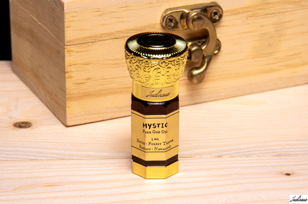 A 3 mL bottle of Mystic OudOil with an ornate gold cap and a gold label, standing on a light wood surface in front of a wooden box with a brass latch.