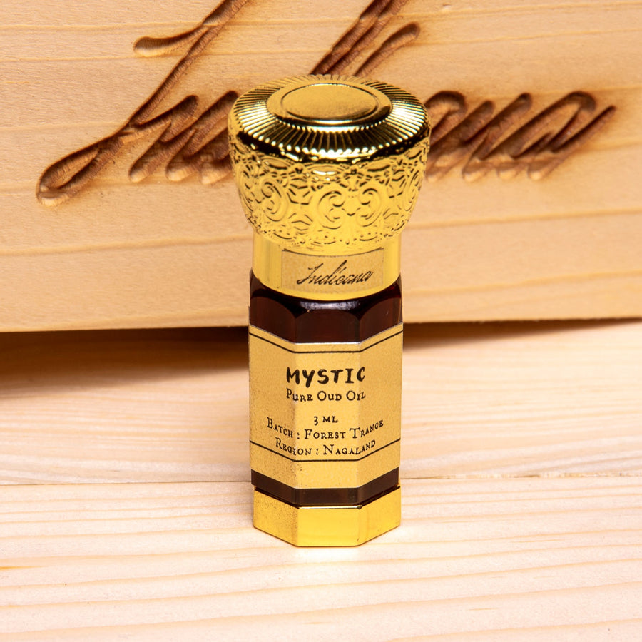 A 3 mL bottle of Mystic OudOil with an ornate gold cap and a gold label, standing on a light wood surface in front of a wooden box with a brass latch.