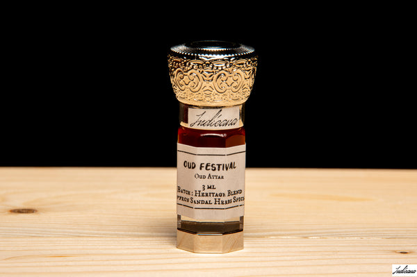 A small glass bottle of Oud Festival Attar, featuring a silver-toned ornate cap and a cream-colored label, set on a light wood surface against a solid black background.