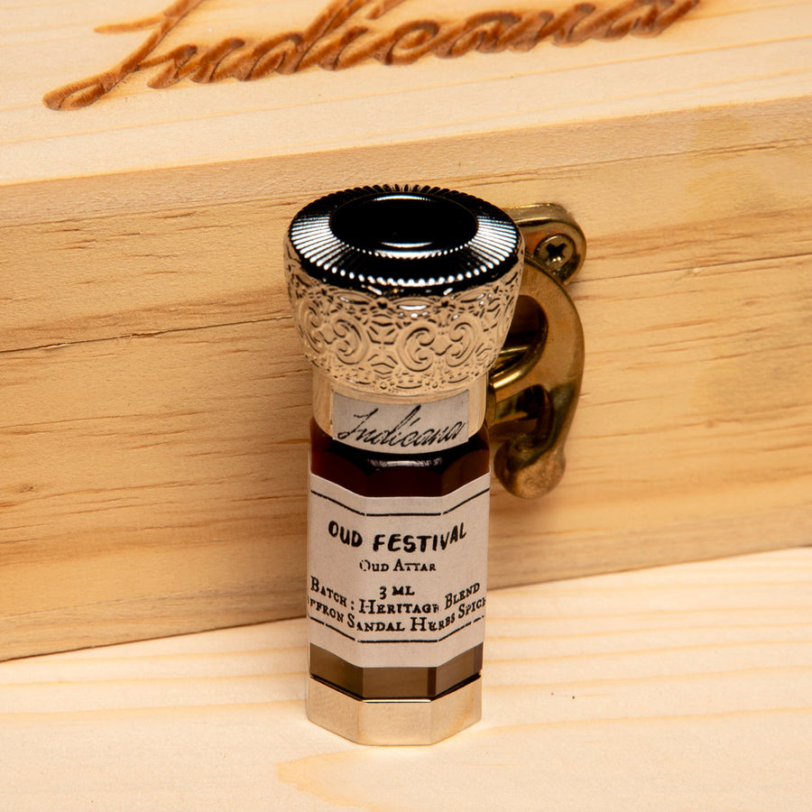 A small glass bottle of Oud Festival Attar, featuring a silver-toned ornate cap and a cream-colored label, set on a light wood surface against a solid black background.