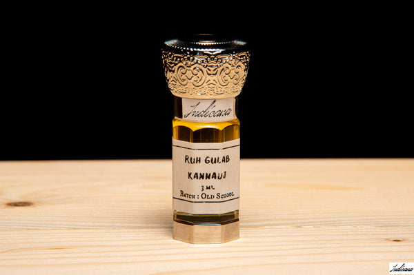 A small glass bottle of Ruh Gulab Kannauj essential oil, featuring a silver cap with an ornate gold filigree pattern and a white label, set on a light wood surface against a solid black background.