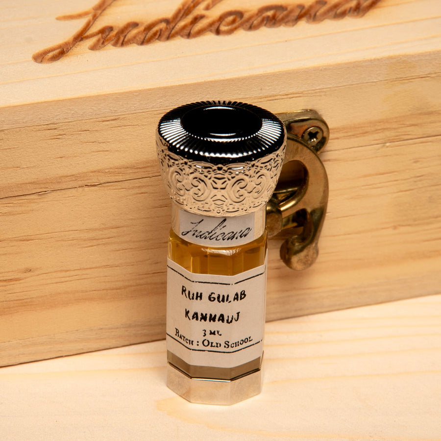A small glass bottle of Ruh Gulab Kannauj essential oil, featuring a silver cap with an ornate gold filigree pattern and a white label, set on a light wood surface against a solid black background.