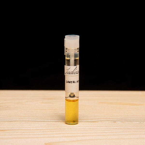 A small glass vial containing a high quality essential oil, standing upright on a light wood surface against a solid black background. The vial has a white label with black text and a white cap.