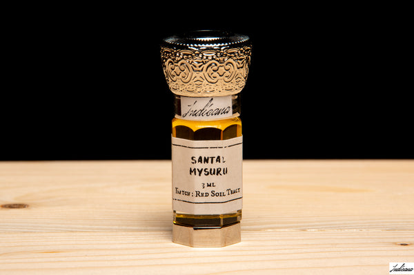 A small glass vial of Santal Mysuru fragrance oil with a white label and ornate silver cap, standing on a light wood surface against a black background.