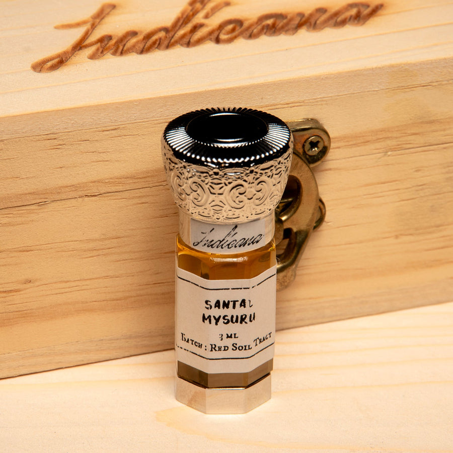 A small glass vial of Santal Mysuru fragrance oil with a white label and ornate silver cap, standing on a light wood surface against a black background.