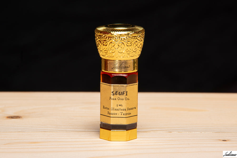 A small ornate perfume bottle with a gold cap and gold label stands in front of an open wooden presentation box on a light wood surface. The label reads 'SEUFI Pour Our Co.