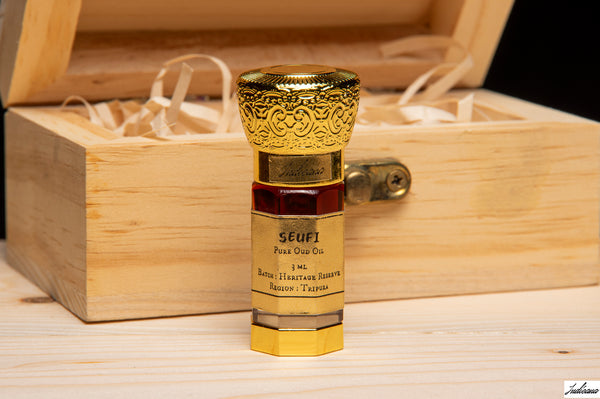 A small ornate perfume bottle with a gold cap and gold label stands in front of an open wooden presentation box on a light wood surface. The label reads 'SEUFI Pour Our Co.