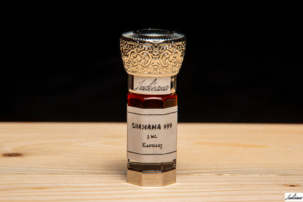 A small glass bottle of Shamama 999 with a white label and ornate silver cap, standing on a light wood surface against a black background.