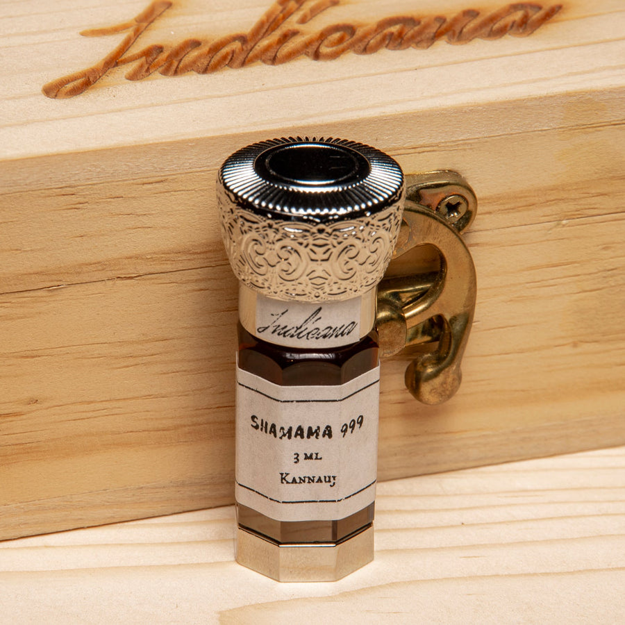 A small glass bottle of Shamama 999 with a white label and ornate silver cap, standing on a light wood surface against a black background.