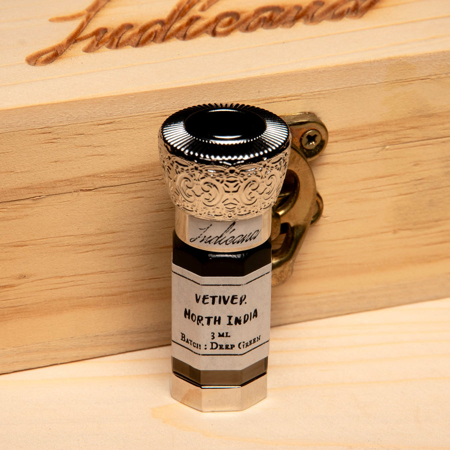 A 3 mL glass bottle of Vetiver North India , deep dark brown shade, featuring a highly detailed ornate gold-toned cap and a white label, set on a light wood surface against a solid black background.